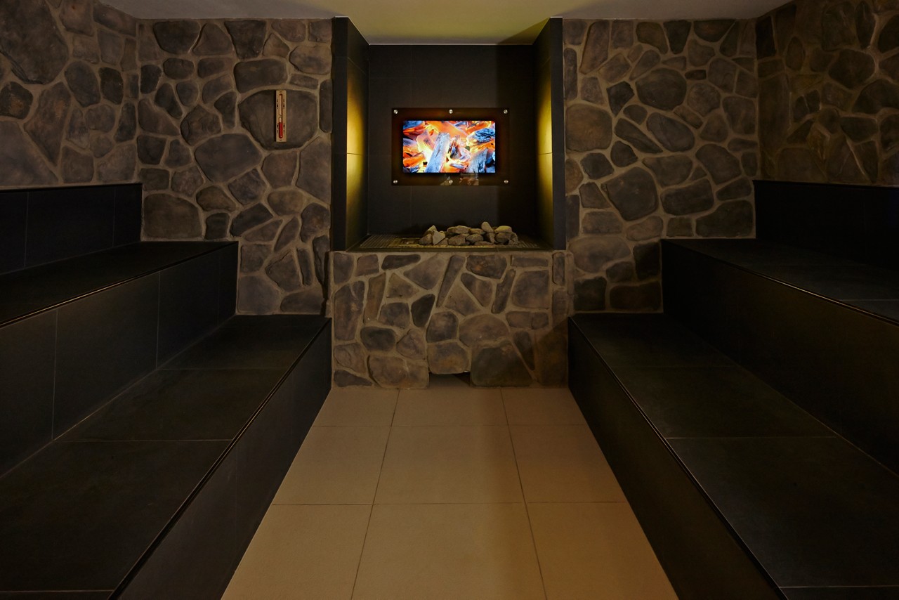 Sauna benches flank a central stone heater; a small wall-mounted screen displays flames, illuminating the dim, stone-walled spa room with a tiled floor.